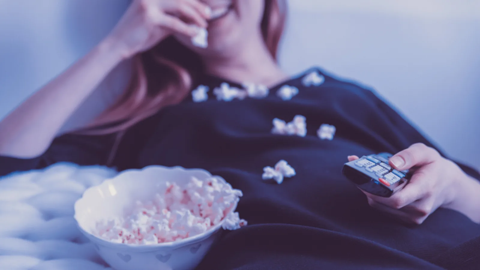 A woman with popcorn holding a TV remote at home.