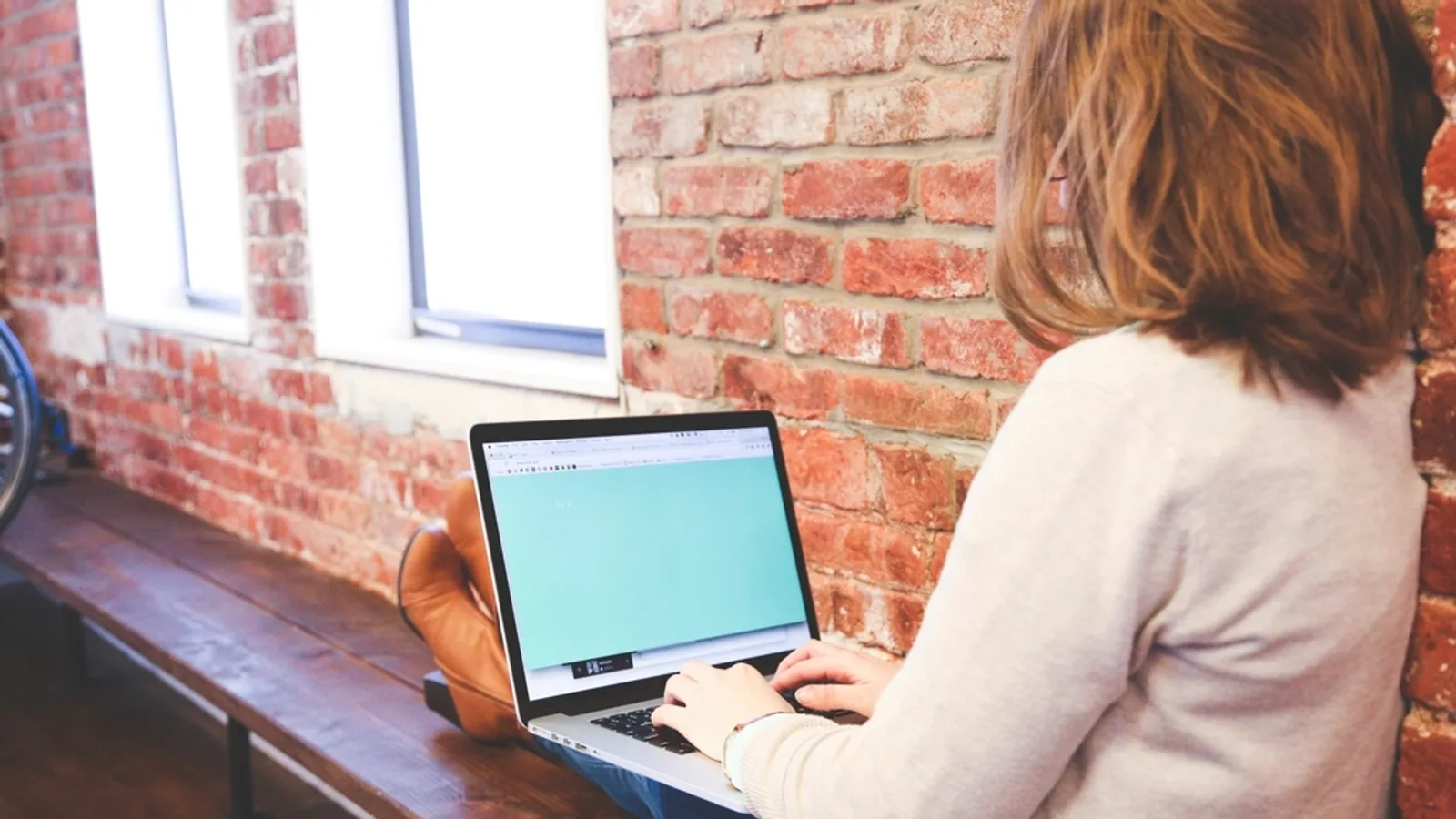 A marketer working on a laptop at a desk by a brick wall.