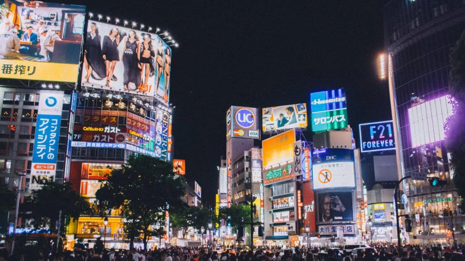 Illuminated LED advertising screens above a busy city crossing at night.